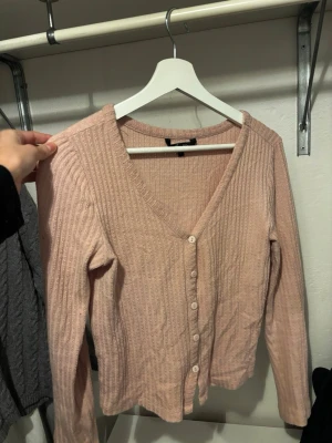 Ljusrosa ribbad kofta med knappar - Söt ljusrosa kofta med ribbad struktur och v-ringning. Koftan har långa ärmar och stängs med vita knappar framtill. Materialet ser mjukt och stretchigt ut, perfekt för lager på lager. Enkel och stilren design som passar till mycket.