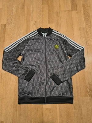Svart Adidas med mönster - Säljer en svart Adidas med ikoniskt mönster och vita ränder längs ärmarna. Jackan har dragkedja framtill, ribbade muddar och gult Adidas-tryck på bröstet. Perfekt för dig som gillar sportig och trendig stil. Stl 176