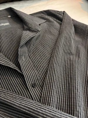 Randig skjorta från Dan Ryan XXL - Snygg svart skjorta från Dan Ryan i storlek XXL med tunna vita ränder. Klassisk krage och knäppning framtill. Skjortan är tillverkad i ett lätt och bekvämt material som passar perfekt till många olika stilar.