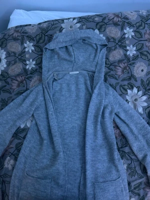 Grå stickad hoodiekofta KappAhl - Mysig grå stickad kofta med huva från KappAhl. Koftan har långa ärmar, öppet framtill och två fickor. Perfekt för lager på lager och chill dagar. Enkel och stilren design som passar till det mesta.