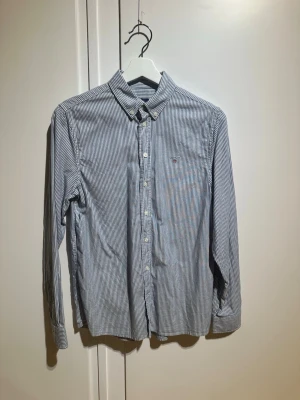 Randig skjorta från Gant - Klassisk blåvit randig skjorta från Gant med lång ärm och knappar framtill. Skjortan har button-down krage och liten broderad logga på bröstet. Tillverkad i mjuk bomull för en bekväm känsla och stilren look. Tveka inte att skriva vid frågor eller funderingar 