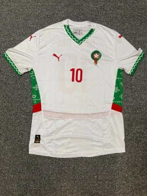 Marocko fotbollströja brahim Diaz  - Snygg vit Marocko fotbollströja från Puma med gröna och röda detaljer. Nummer 10 och namnet BRAHIM på ryggen. Tröjan har gröna mönstrade ärmslut och V-ringning, samt Marockos landslagsemblem på bröstet. Det står XL men den passar som M