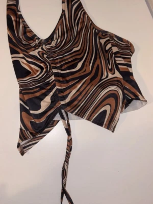 Brun mönstrad croppad linnetopp - Säljer en croppad linnetopp med brunt, svart och beige abstrakt mönster. Toppen har smala axelband, snörning framtill och är tillverkad i ett stretchigt syntetmaterial. Perfekt för dig som gillar trendiga och unika plagg.