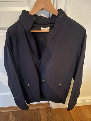 Mörkblå Zadig jacka - Jacka från Zadig. Jackan har två framfickor med tryckknappar och är tillverkad i ett lätt syntetmaterial. Perfekt för blåsiga och regniga dagar och enkel att matcha med olika outfits.  En liten fläck som knappt syns