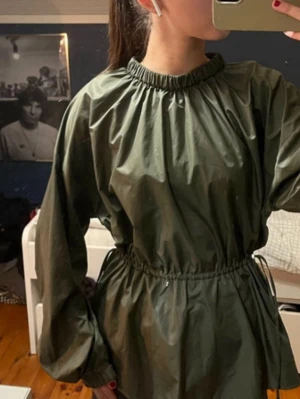 Mörkgrön vindjacka med resårmidja - Snygg mörkgrön vindjacka med resår i midjan och vid halsen. Jackan har lång ärm och en loose passform, perfekt för lager-på-lager. Tillverkad i ett syntetmaterial som står emot vind. Enkel och stilren design utan synliga loggor.