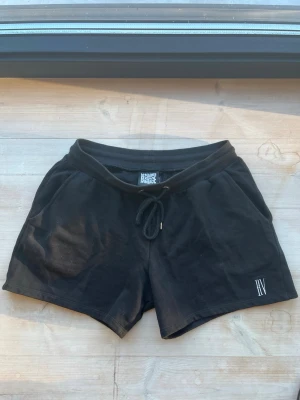 Svarta shorts från Reclaimed Vintage strl M - Svarta shorts från Reclaimed Vintage med snörning i midjan och diskret logga på benet. Tillverkade i mjuk bomull, perfekta för avslappnade dagar. Klassisk passform och enkel design som funkar till det mesta.