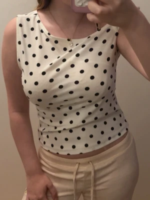 Vit prickig topp - Snygg vit ärmlös topp med svarta prickar och rund halsringning. Toppen har en figurnära passform och är tillverkad i ett mjukt, stretchigt material som känns bekvämt mot huden. Perfekt för dig som gillar en lekfull och trendig stil.