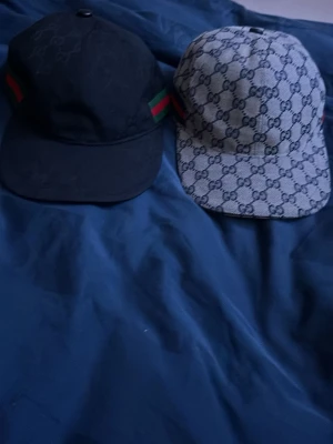 Gucci kepsar med GG-mönster - Två snygga Gucci kepsar med klassiskt GG-mönster. Ena kepsen är svart med gröna och röda ränder på sidorna, den andra är ljusgrå med mörkare GG-loggor. Justerbar rem baktill i läderdetalj. Perfekt accessoar för att lyfta din stil. Det är 900 styck och om du vill köpa båda får du de för 1500