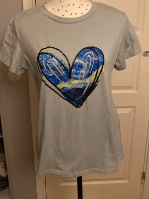 Ljusgrå t-shirt med blått hjärta - Snygg ljusgrå t-shirt med ett stort hjärta i blått, vitt och svart på bröstet. T-shirten har korta ärmar och en klassisk rund halsringning. Perfekt för dig som gillar konstnärliga tryck och vill ha en unik look.