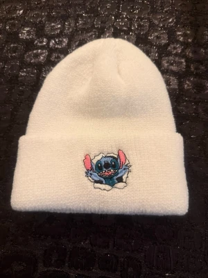 Vit mössa med Stitch-brodyr - Säljer en vit stickad mössa med uppvikt kant och en broderad Stitch från Lilo & Stitch på framsidan. Mössan är mjuk och har en cool, ungdomlig vibe med sin tecknade detalj. Perfekt för dig som gillar Disney och vill ha något unikt till vintern. Mössan är helt ny och aldrig använd. 