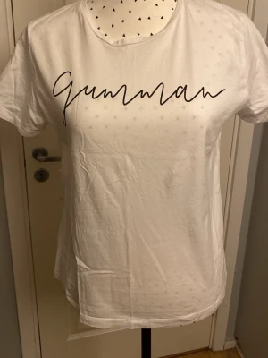 Vit t-shirt med texten Gumman - Vit t-shirt med svart texten 'Gumman' i snygg skrivstil över bröstet. Klassisk rund halsringning och korta ärmar. Perfekt basic-plagg med en personlig touch, tillverkad i mjuk bomull för skön känsla.