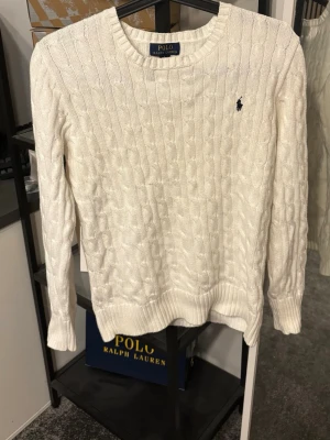 Vit kabelstickad tröja Polo Ralph Lauren - Kabelstickad vit tröja från Polo Ralph Lauren med rund halsringning och broderad logga på bröstet. Tröjan har långa ärmar och ribbade muddar vid ärmslut och nederkant. Perfekt för en stilren och klassisk look.