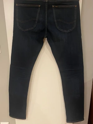 Mörkblå slim jeans från Lee - Snygga mörkblå slim jeans från Lee med klassiska bakfickor och bruna kontrastsömmar. Jeansen har en smal passform och är tillverkade i mjukt denimtyg med Lee-logga på bakfickan och läderpatch i midjan. Hör av er vid minsta fråga 😍😍