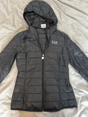 Svart dunjacka från EA7 Emporio Armani - Snygg svart dunjacka från EA7 Emporio Armani med huva och dragkedja framtill. Jackan har quiltad design och logga på bröstet. Perfekt för kyliga dagar och enkel att matcha med olika outfits.