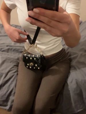 Svart quiltad axelväska med pärlor - Snygg svart quiltad axelväska med pärldetaljer och guldfärgad kedja. Väskan har en elegant vridlåsdetalj framtill och justerbar axelrem. Perfekt för att ge en touch av glamour till din outfit.