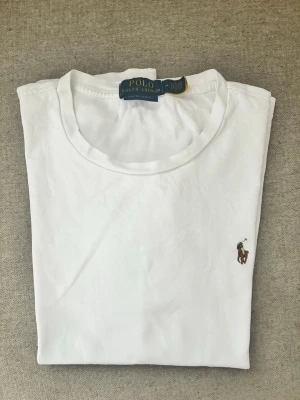 Vit t-shirt från Polo Ralph Lauren - Klassisk vit t-shirt från Polo Ralph Lauren med rund hals och den ikoniska broderade polospelaren på bröstet. Tillverkad i mjuk bomull för en skön och avslappnad look. Perfekt basplagg till garderoben.
