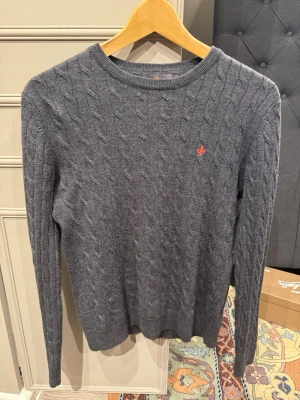 Grå kabelstickad tröja med brodyr - Mysig grå kabelstickad tröja med rund halsringning och en liten orange broderad detalj på bröstet. Tröjan har långa ärmar och ribbade muddar. Perfekt för dig som gillar klassisk stil med en twist.