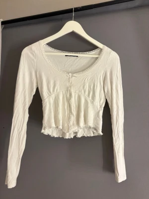 Vit croppad långärmad topp med spets - Söt vit croppad topp med långa ärmar och spetsdetaljer längs urringningen och framtill. Toppen har en liten rosett framtill och volangkant nedtill för en romantisk vibe. Perfekt att styla med jeans eller kjol.