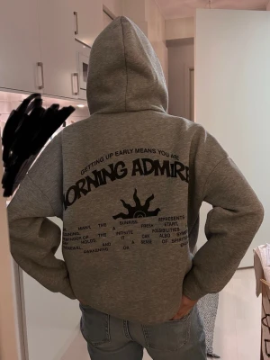 Grå hoodie med tryck 'Morning Admire' - Säljer en grå hoodie med svart tryck på ryggen där det står 'Morning Admire' och en solgrafik. På framsidan finns mindre texttryck. Hoodien har huva och är i mjukt material, perfekt för en avslappnad stil. Tröjan är köpt från SHEIN , och har luddiga ställen men inte jätte farligt. Passformen är mellan xxs och xs.