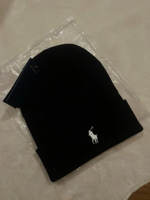 Svart mössa från Polo Ralph Lauren - Snygg svart mössa från Polo Ralph Lauren med klassisk vit broderad logga framtill. Enkel och stilren design med uppvikt kant. Perfekt accessoar för kalla dagar och passar till det mesta. Materialet känns mjukt och bekvämt.