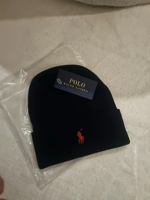 Marinblå mössa från Polo Ralph Lauren - Snygg marinblå mössa från Polo Ralph Lauren med klassisk uppvikt kant och röd broderad logga framtill. Perfekt accessoar för att hålla värmen och samtidigt se stilren ut. Mjuk och stretchig kvalitet som sitter skönt på huvudet.