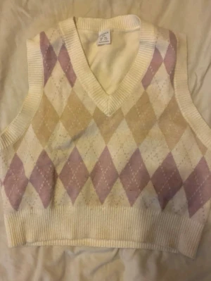 Kritvit argyle väst med pastellrutor - Snygg stickad väst i kritvitt med klassiskt argylemönster i pastellila och beige. Djup V-ringning och bred ribbad kant nedtill och vid ärmhålen. Perfekt för lager-på-lager och ger en retrovibe till din outfit.