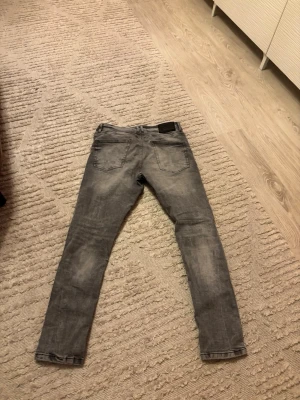 Grå slitna skinny jeans - Säljer ett par grå skinny jeans med slitningar på framsidan. Jeansen har klassisk femficksdesign och är tillverkade i stretchigt denimtyg för en tajt passform. Perfekta för dig som gillar en edgy och trendig stil.