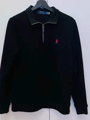Ralph Lauren Quarter Zip (Copy) - Black long-sleeved sweater from Ralph Lauren with half zip and classic collar. The sweater has a red embroidered logo on the chest and is made of soft material that feels nice against the skin. Perfect for a stylish and relaxed look. (Copy)