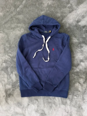 Blå hoodie från Polo Ralph Lauren - Snygg blå hoodie från Polo Ralph Lauren i slim fit-modell. Hoodien har vit dragsko, klassisk huva och den ikoniska röda loggan broderad på bröstet. Tillverkad i mjuk bomullsblandning och har ribbade muddar vid ärmar och nederkant.