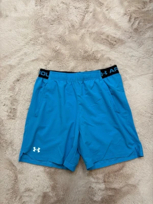 Under Armour - Snygga blå träningsshorts från Under Armour med elastisk midja och logga på benet. Shortsen har praktiska sidofickor och svart resår med märke upptill. Perfekta för sport eller gympass.