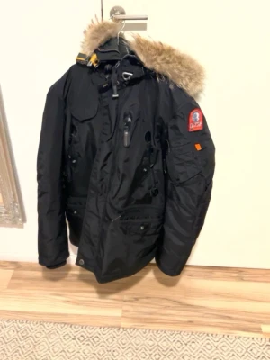 Svart Parajumpers parkas  - Svart parkajacka från Parajumpers med avtagbar päls på huvan, flera fickor och gul detalj vid kragen. Jackan har dragkedja och knappar framtill samt patch på ärmen. Perfekt för kalla dagar och riktigt snygg streetstil.