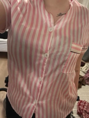 Rosa och vitrandig pyjamaströja - Kortärmad pyjamaströja med rosa och vita vertikala ränder. Knäppning framtill med vita knappar och en bröstficka med svart detalj. Skön och luftig modell, perfekt för mysiga kvällar hemma.