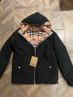 Burberry Jacka - Svart vändbar jacka från Burberry. Den ena sidan är svart och den andra har det ikoniska Burberry-rutmönstret. Jackan har en huva, dragkedja fram och två fickor. Perfekt för höst och vår. Denna var för stor för mig så typ aldrig använd.