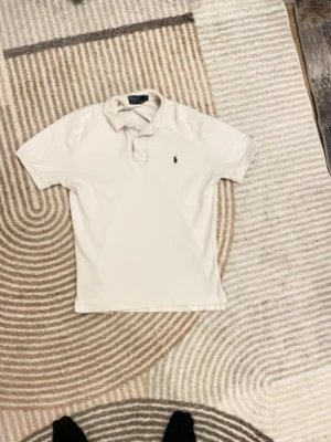 Söker byte  - Klassisk vit pikétröja från Polo Ralph Lauren med blå logga sopatchad på bröstet. Tröjan har korta ärmar, krage och knappar vid halsen. Perfekt till jeans eller shorts för en clean och stilren look.