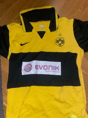 Borussia Dortmund matchtröja Nike storlek M - Gul och svart Borussia Dortmund matchtröja från Nike med klubbmärke och sponsortryck från Evonik Industries. Klassisk krage med svarta detaljer och broderad Nike-logga. Perfekt för fotbollsfans som vill sticka ut på läktaren eller planen.