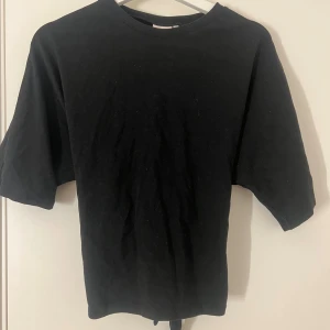 Svart croppad topp med knyt - Svart croppad topp med korta ärmar och snygg knytning i ryggen. Enkel och stilren design med rund halsringning. Perfekt för att skapa en chill och trendig look. Mjuk och skön bomullskänsla som gör den bekväm att bära hela dagen.