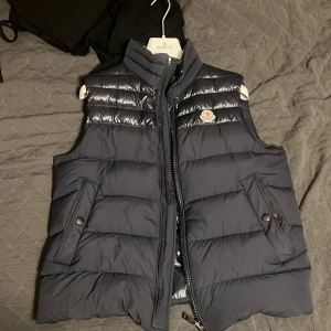 Mörkblå dunväst från Moncler - Snygg mörkblå dunväst från Moncler med quiltad design och hög krage. Västen har två fickor med tryckknappar, dragkedja framtill och Moncler-logga på bröstet. Insidan har ett coolt serietryck.  Västen är fortfarande i så gott skick som när jag köpte den från butik, inga brister och säljer för att jag inte använder den längre. 