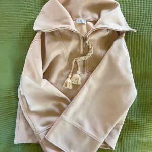 Claudie Pierlot quarter zip-tröja - Ljust puderrosa tröja från Claudie Pierlot med quarter zip och flätad snörning med tofsar. Tröjan har hög krage och vida ärmar. Passar M/L men även S som over size