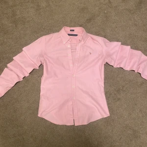 Rosa skjorta från Ralph Lauren - Snygg rosa skjorta från Ralph Lauren i slim fit med klassisk krage och broderad logga på bröstet. Skjortan har långa ärmar och knappar framtill. Perfekt för dig som gillar stilrena och fräscha plagg med en ikonisk touch.