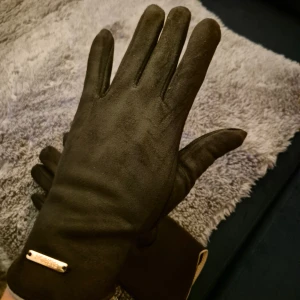 Svarta mockahandskar med detalj - Snygga svarta handskar i mocka med en stilren metallbricka där det står 'FASHION' vid handleden. Handskarna har en klassisk, slimmad passform och är perfekta för kyliga dagar. Insidan är mjuk och de har en diskret beige kant längst ner.