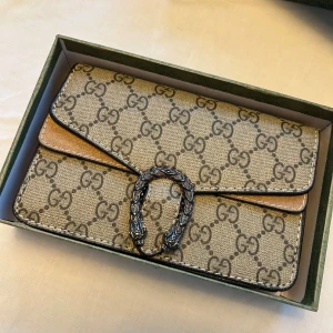 Gucci kuvertväska med GG-mönster - Snygg kuvertväska Gucci i beige och brunt med klassiskt GG-mönster över hela väskan. Väskan har en dekorativ metallspänne framtill och levereras i originalbox och dustbag. Perfekt accessoar för att lyfta din outfit.