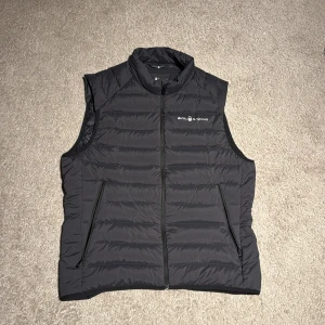 Svart dunväst från Sail Racing - Svart dunväst från Sail Racing med quiltad design och hög krage. Västen har två dragkedjeförsedda fickor på framsidan och logotyp på bröstet samt ryggen. Perfekt att slänga över en hoodie för extra värme. Snygg och stilren look.