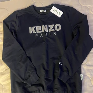 Ny Svart sweatshirt från Kenzo Paris - Svart sweatshirt från Kenzo Paris med broderad logga framtill i vitt och grått. Tröjan har rund hals, ribbade muddar och är tillverkad i mjuk bomull. Klassisk och stilren design med Kenzo-detaljer på ärm och nederkant.