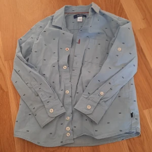 Ljusblå skjorta från Okaidi 104cm - Ljusblå skjorta från Okaidi med små mörkblå rektangulära mönster, klassisk krage och vita knappar. Skjortan har lång ärm och bröstficka, perfekt för en stilren look. Tillverkad i mjuk bomull som känns skön mot huden.