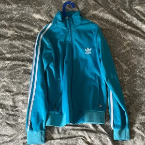 Turkos Adidas zip up strl M - Snygg turkos zip up från Adidas med klassiska vita ränder längs ärmarna och logga på bröstet. Jackan har hög krage, dragkedja framtill och två fickor med zip. Perfekt för dig som gillar sportig stil och vill sticka ut. Skriv vi mer funderingar😇👍🏼