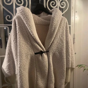 Beige teddyjacka med huva - Mysig beige teddyjacka med stor huva och breda ärmar. Jackan har ett snyggt träknappsspänne framtill och ribbstickade muddar vid ärmsluten. Perfekt för kalla dagar när du vill ha en chill och varm look.