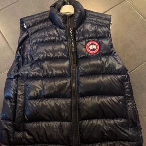Mörkblå dunväst från Canada Goose - Snygg mörkblå dunväst från Canada Goose med klassisk logga på bröstet och hög krage. Västen har quiltad design, dragkedja framtill och är perfekt för lager-på-lager. Materialet är glansigt och känns riktigt lyxigt.
