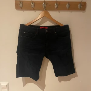 Svarta jeansshorts från Hugo Boss - Svarta jeansshorts från Hugo Boss med klassisk femficksdesign och knappgylf. Snygg, stilren look med mörk tvätt och diskreta sömmar. Perfekta för dig som gillar en enkel men trendig stil. Passar till allt och har en bekväm midja.