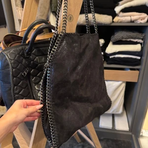 Svart axelväska med kedjedetaljer - Cool svart axelväska liknar Stella McCartney i mocka-look med silverfärgade kedjor längs kanterna och som axelrem. Väskan har ett snyggt mönstrat innerfoder och dragkedjefack inuti. Perfekt för dig som gillar edgy och trendig stil.