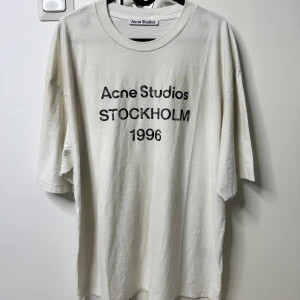 Vit Acne Studios logo t-shirt med Stockholm 1996 tryck - Vit t-shirt från Acne Studios med svart texttryck 'Acne Studios STOCKHOLM 1996' på bröstet. Klassisk rund halsringning och avslappnad passform. Tillverkad i mjuk bomull, perfekt för en clean och stilren look.